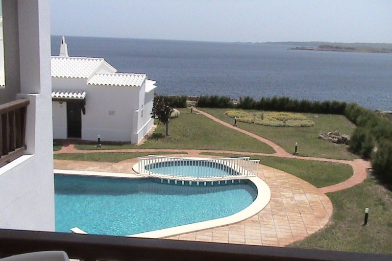 Vista de la piscina del complejo de apartamentos Rocas Marinas y la costa de Menorca de fondo.